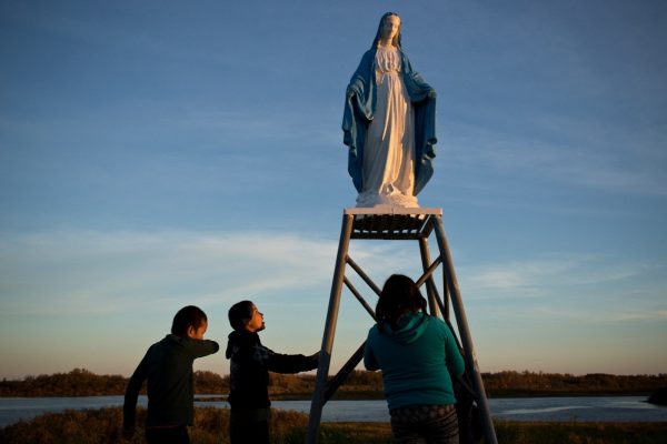 Attawapiskat, Ontario, Canada. Young people play at a statue of the Virgin Mary near the St. Francois-xavier Catholic Church on the shores of the Attawapiskat river. The community has a complicated history with the Catholic church with many of its members subjected to horrible abuses while in the Catholic run residential school system.

Attawapiskat is an isolated First Nation community  located in northern Ontario, Canada, at the mouth of the Attawapiskat River on James Bay. On April 9, 2016, the community of approximately 2000 people declared a state of emergency after being overwhelmed with attempted suicides, over 100 attempts in a ten month period. October 7, 2016. David Maurice Smith/Oculi.